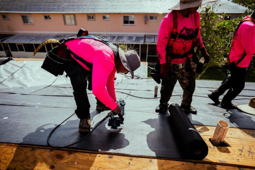 A worker using a nail gun secures underlayment on a wooden roof.