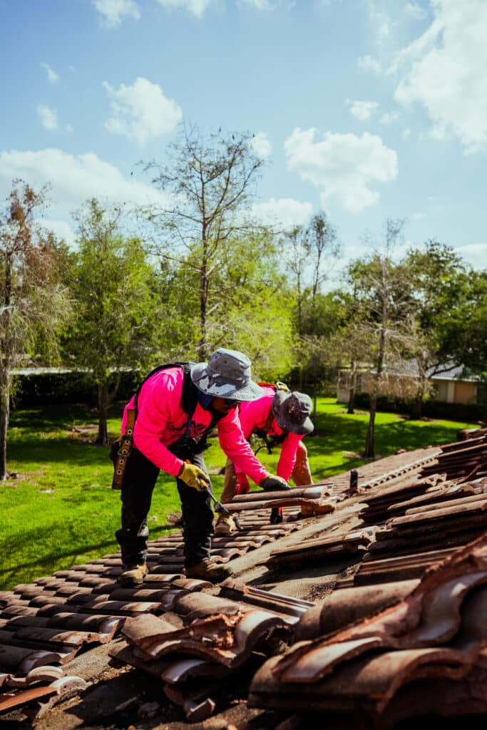Two roofers in safety gear install dark tiles on a residential roof.